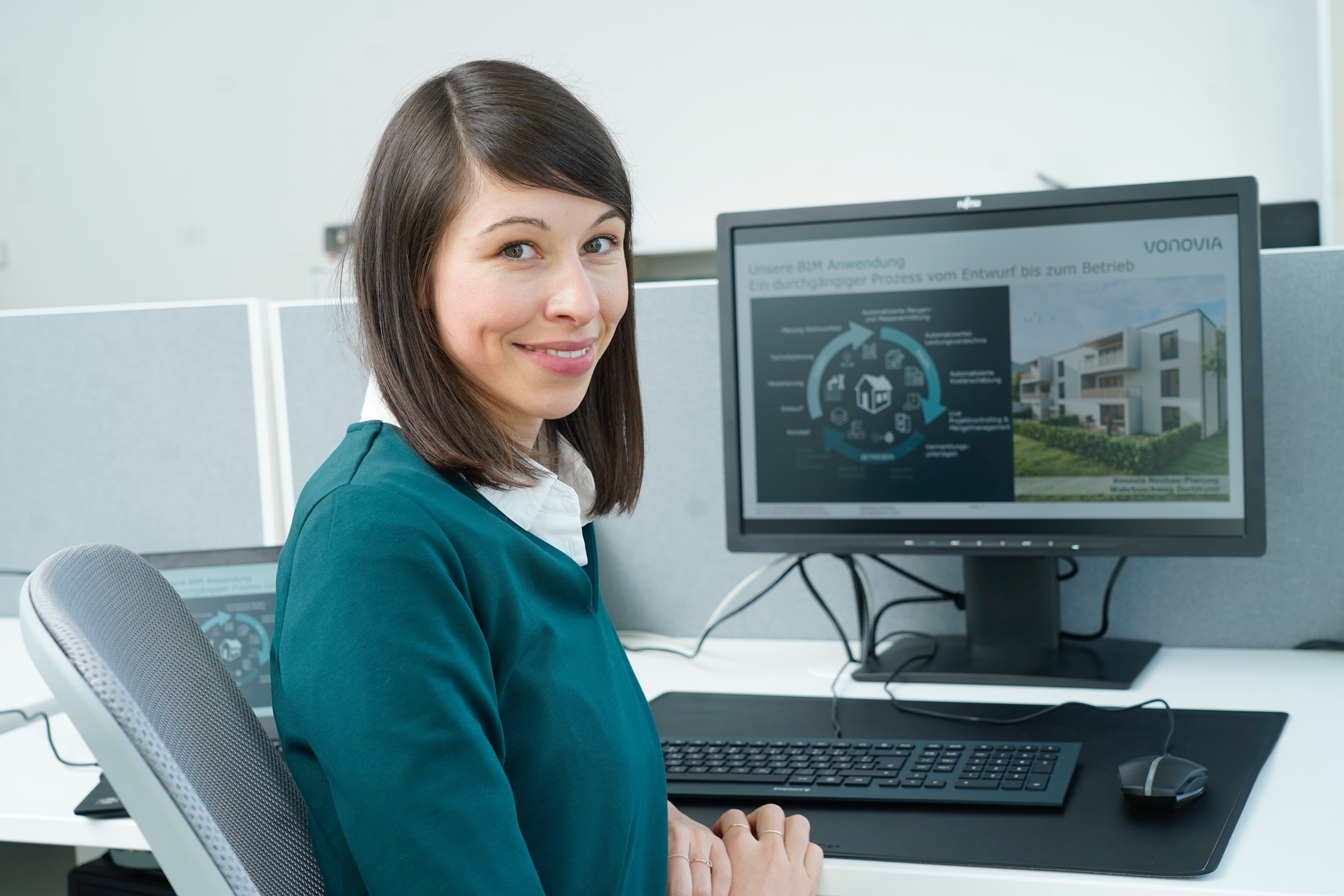 Frau mit braunen Haare sitzt seitlich vor einem Computerbildschirm, auf welchen man ein Flowchart der Vonovia leicht verschwommen erkennen kann, und lächelt in die Kamera. Maus und Tastatur liegen auf einem großen Mauspad auf dem grauen Schreibtisch vor ihr.